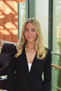 Blonde woman in black blazer in front of windows.