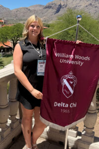 Blonde woman in black dress holding a banner that says Delta Chi with an Alpha Phi crest.